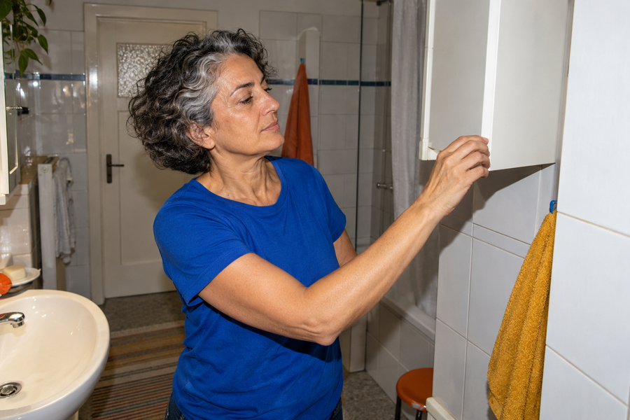 Eine Frau mit grauen Locken und blauem T-Shirt schließt einen Wandschrank im Badezimmer.